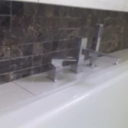 Bateria umywalkowa o kwadratowym designie zamontowana na białym blacie, tło stanowi ściana wyłożona mozaiką z ciemnego marmuru.