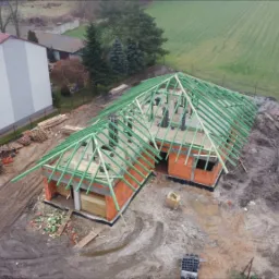 Zdjęcie z lotu ptaka prezentuje budowę domu z czerwonej cegły z zieloną konstrukcją dachu, obok stoi trzypiętrowy budynek mieszkalny z balkonami, a w tle widać pole i drzewa.