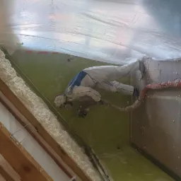 Pracownik w kombinezonie nanosi piankę PUR na poddasze z drewnianą konstrukcją. Widoczny wąż i pianka w narożnikach. Ochrona folią na suficie.