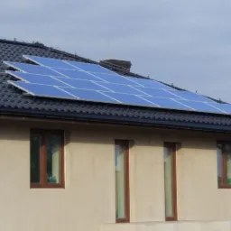 Panel słoneczny na dachu domu jednorodzinnego z ciemną dachówką, widoczne trzy okna na ścianie budynku, pochmurne niebo.