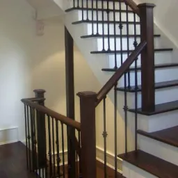 Klatka schodowa z ciemnego drewna z czarnymi metalowymi balustradami, widok z dołu schodów, jasne ściany.