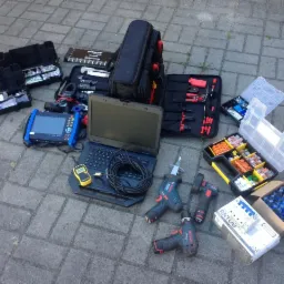 Zestaw narzędzi i sprzętu do instalacji: laptop, tester kabli, wkrętarki Bosch, skrzynki z elementami złącznymi, torba narzędziowa, ułożone na kostce brukowej.