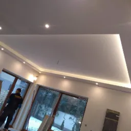 Podwieszany sufit z oświetleniem LED punktowym i liniowym w nowo wykończonym wnętrzu, widoczny pracownik na drabinie.