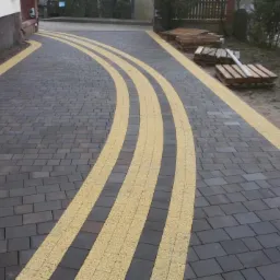 Ukończona nawierzchnia z ciemnoszarej kostki brukowej z trzema zakrzywionymi pasami żółtej kostki, palety z kostką w tle.