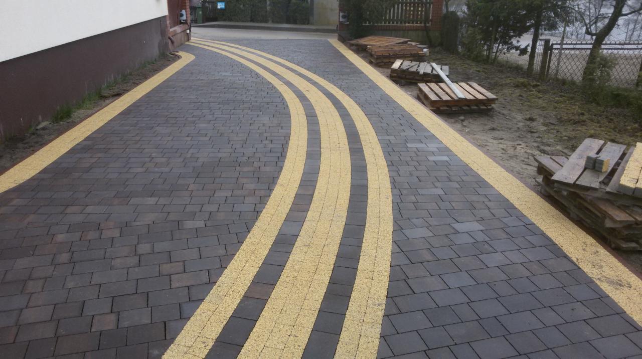 Ukończona nawierzchnia z ciemnoszarej kostki brukowej z trzema zakrzywionymi pasami żółtej kostki, palety z kostką w tle.