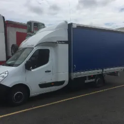 Biały samoch&oacute;d dostawczy z plandeką w kolorze niebieskim, zaparkowany na parkingu z ż&oacute;łtymi liniami, obok innych pojazd&oacute;w ciężarowych.