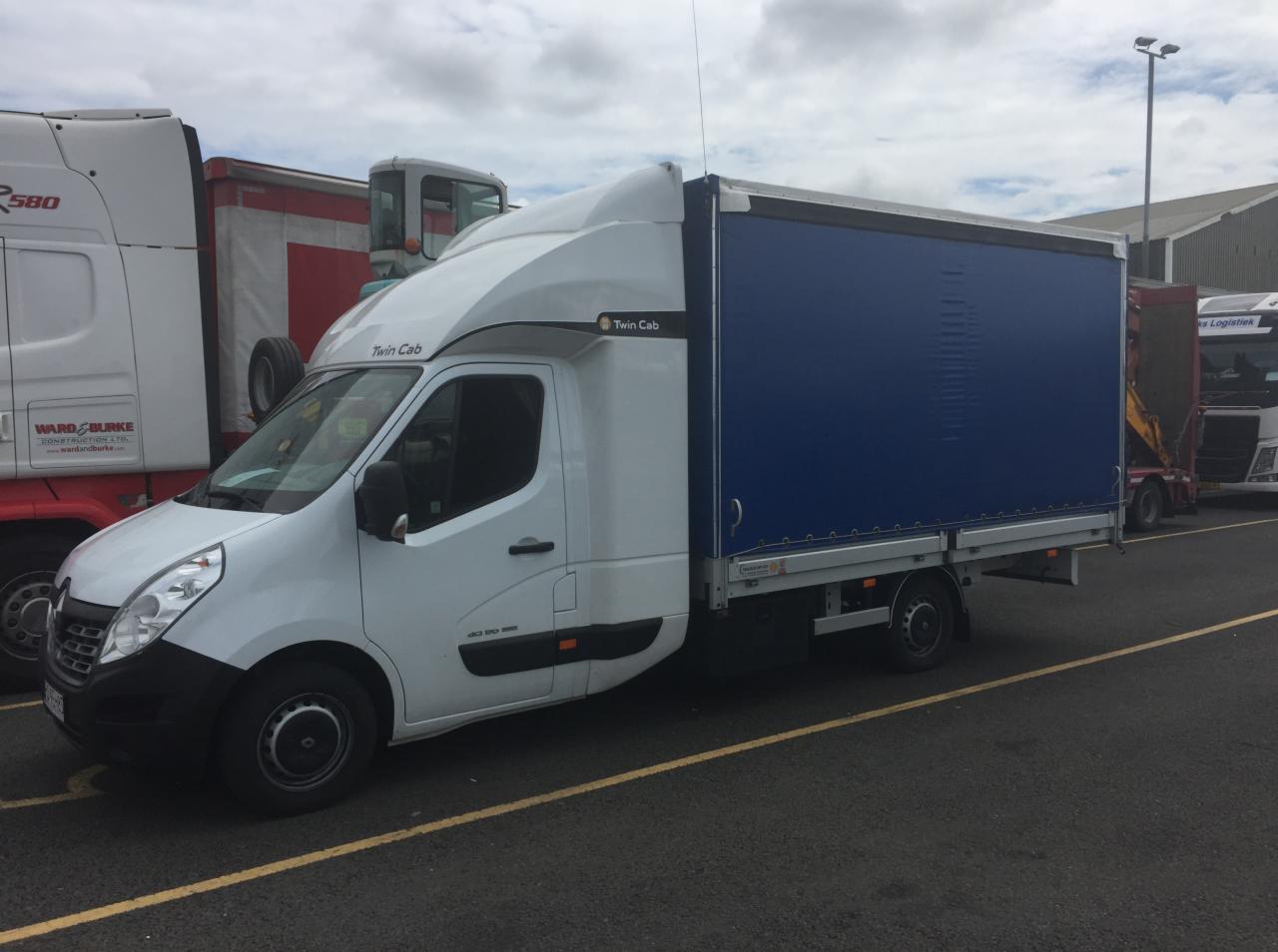 Biały samochód dostawczy z plandeką w kolorze niebieskim, zaparkowany na parkingu z żółtymi liniami, obok innych pojazdów ciężarowych.