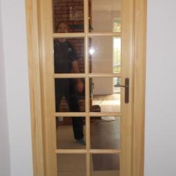 Lumar s.c. Łukasz Gromek Mariusz Zebrowski - Drewniane drzwi wewnętrzne z szybami w szprosach, widoczne odbicie osoby i wnętrze z ceglanym kominkiem. Naturalne wykończenie drewna, metalowa klamka.