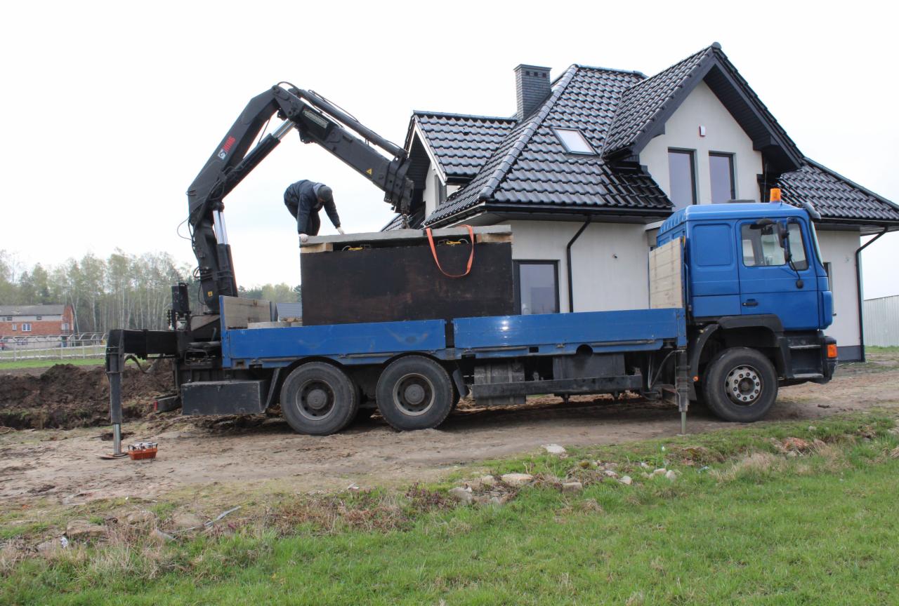 Montaż betonowego zbiornika na deszczówkę za pomocą dźwigu samochodowego na tle domu z czarną dachówką.