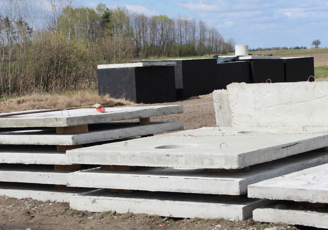 Stos betonowych płyt z okrągłym otworem, z widocznymi metalowymi uchwytami, składowanych na placu budowy w słoneczny dzień, z lasem i polem w tle.