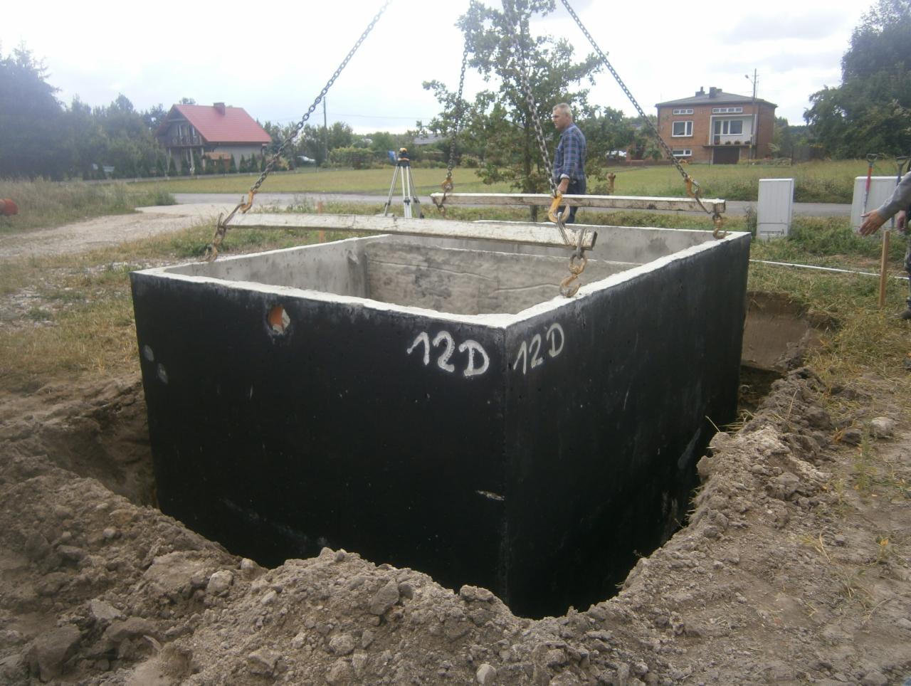 Ciężki, betonowy prefabrykat w kształcie kwadratowej studni, z napisem '12D', opuszczany do wykopu za pomocą łańcuchów, w tle pracownik i domy.
