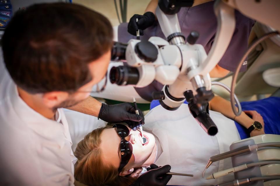 Dentysta w czarnych rękawiczkach przeprowadza zabieg stomatologiczny u pacjentki z osłoną na oczach, używając mikroskopu operacyjnego i narzędzi dentystycznych.
