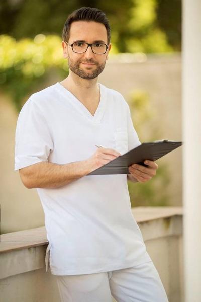 Lekarz w białym uniformie, z okularami, piszący na czarnej podkładce, oparty o betonową ścianę na tle zieleni.