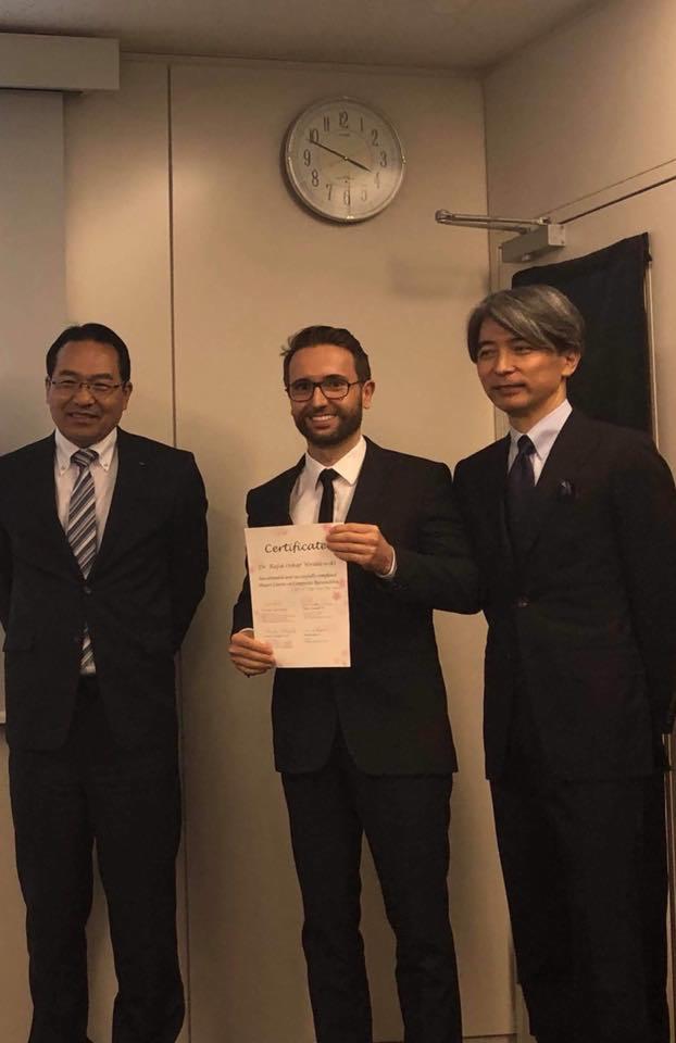 Trzech mężczyzn w garniturach, środkowy trzyma certyfikat w dłoniach, uśmiecha się, na ścianie zegar.