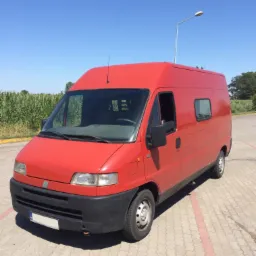 Czerwony, wysoki bus Fiat Ducato zaparkowany na kostce brukowej w słoneczny dzień, z polem kukurydzy i latarnią w tle.