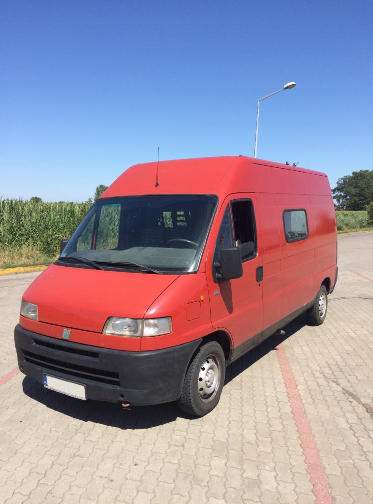 Czerwony, wysoki bus Fiat Ducato zaparkowany na kostce brukowej w słoneczny dzień, z polem kukurydzy i latarnią w tle.