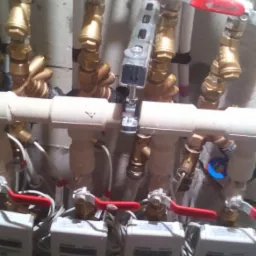 Złożona instalacja wodna z licznymi zaworami z czerwonymi uchwytami, rurami z tworzywa i mosiądzu oraz trzema licznikami wody na dole kadru.