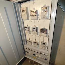 K&M Instalacje elektryczne Krzysztof Mroczko - Otwarta skrzynka z licznikami energii elektrycznej dla mieszkań w Gliwicach. Widoczne liczniki z oznaczeniami mieszkań i zabezpieczenia.