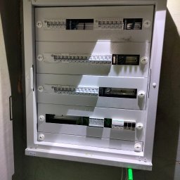 K&M Instalacje elektryczne Krzysztof Mroczko - Szara, otwarta szafa elektryczna marki Eaton z widocznymi bezpiecznikami i przewodami, zamontowana na ścianie. Widoczny fragment przewodu z zieloną końcówką.