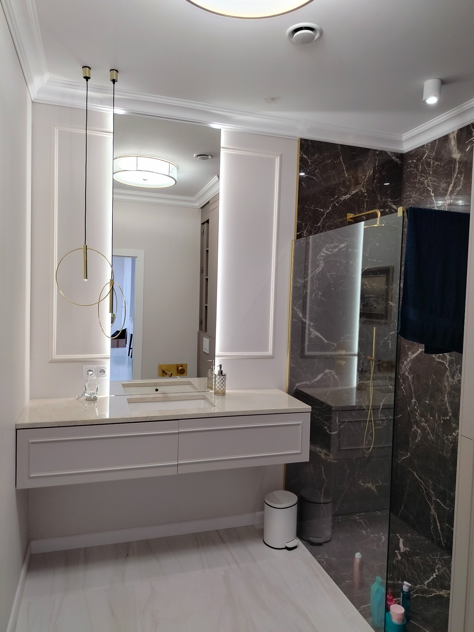 Elegancka łazienka z marmurowymi akcentami, podwieszaną szafką z umywalką i okrągłymi lampami wiszącymi. Widoczna kabina prysznicowa z ciemnym marmurem.