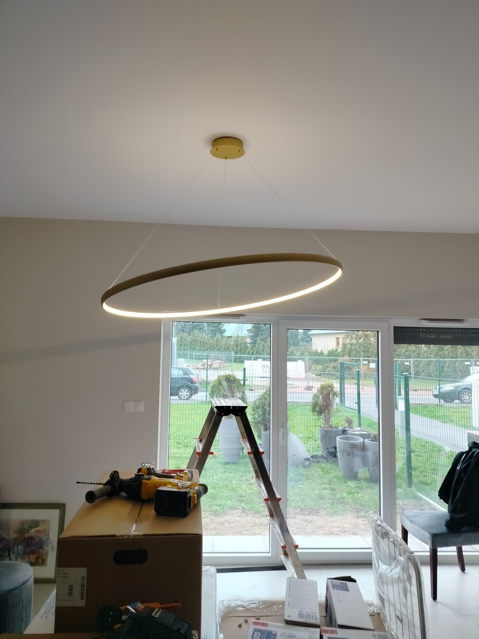 Montaż nowoczesnej, okrągłej lampy LED w jasnym wnętrzu. Widoczna drabina, narzędzia i kartony, sugerujące trwający proces instalacji.