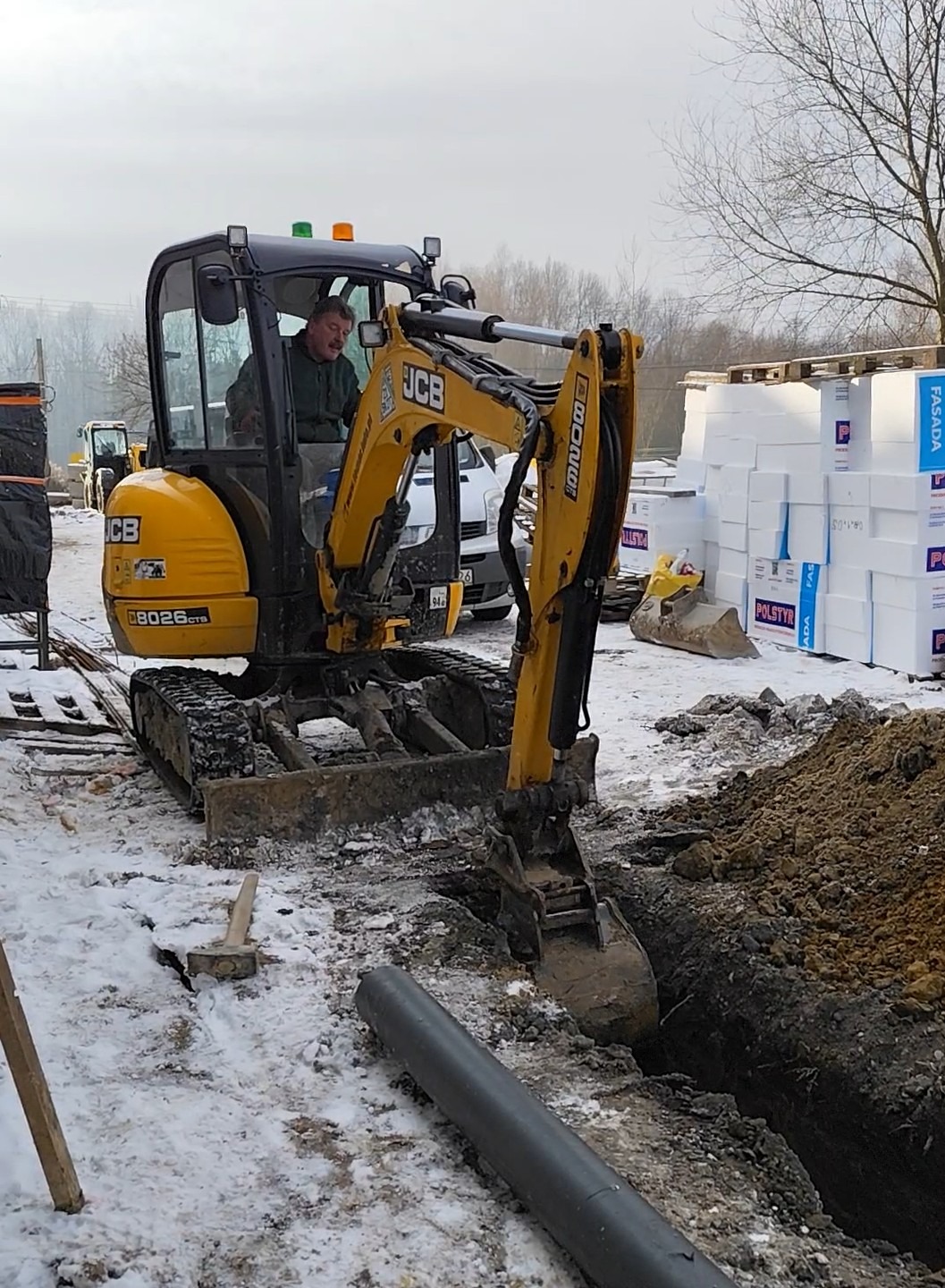 Żółta koparka JCB wykopuje rów w zasypanym śniegiem terenie budowy, z widoczną rurą i stertą styropianu w tle. Operator w kabinie. Prace ziemne w zimowych warunkach.
