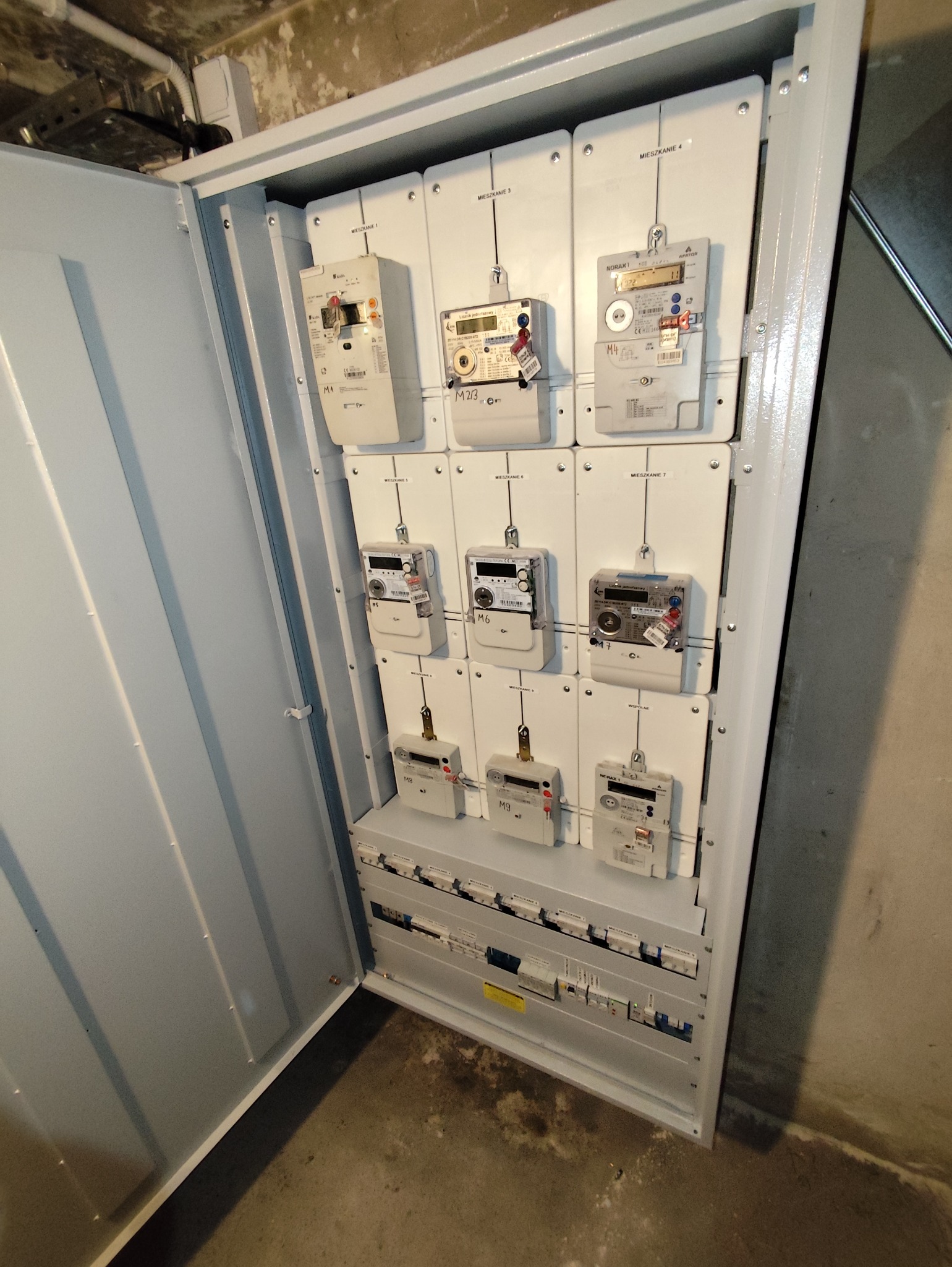 Otwarta skrzynka z licznikami energii elektrycznej dla mieszkań w Gliwicach. Widoczne liczniki z oznaczeniami mieszkań i zabezpieczenia.