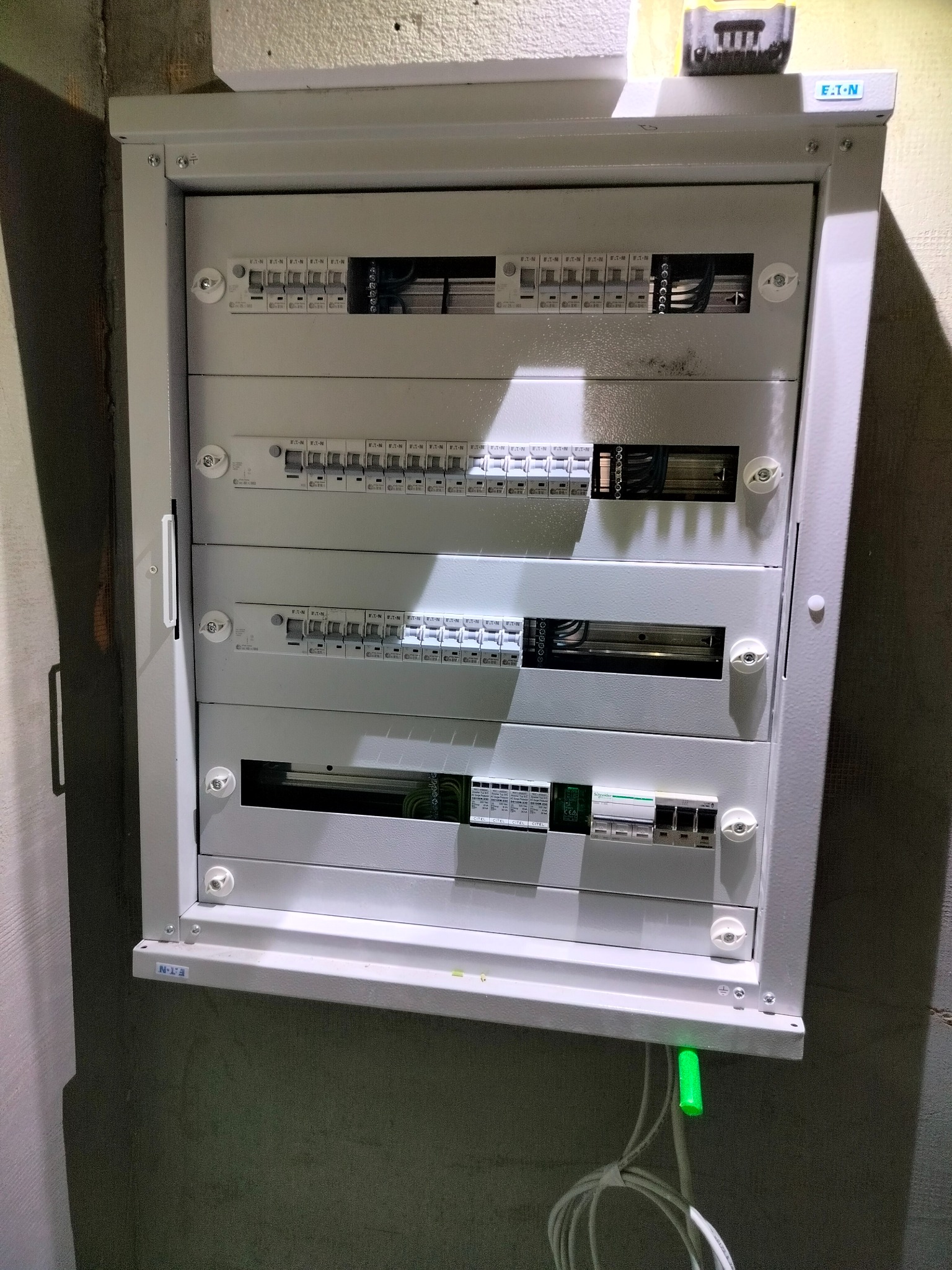 Szara, otwarta szafa elektryczna marki Eaton z widocznymi bezpiecznikami i przewodami, zamontowana na ścianie. Widoczny fragment przewodu z zieloną końcówką.