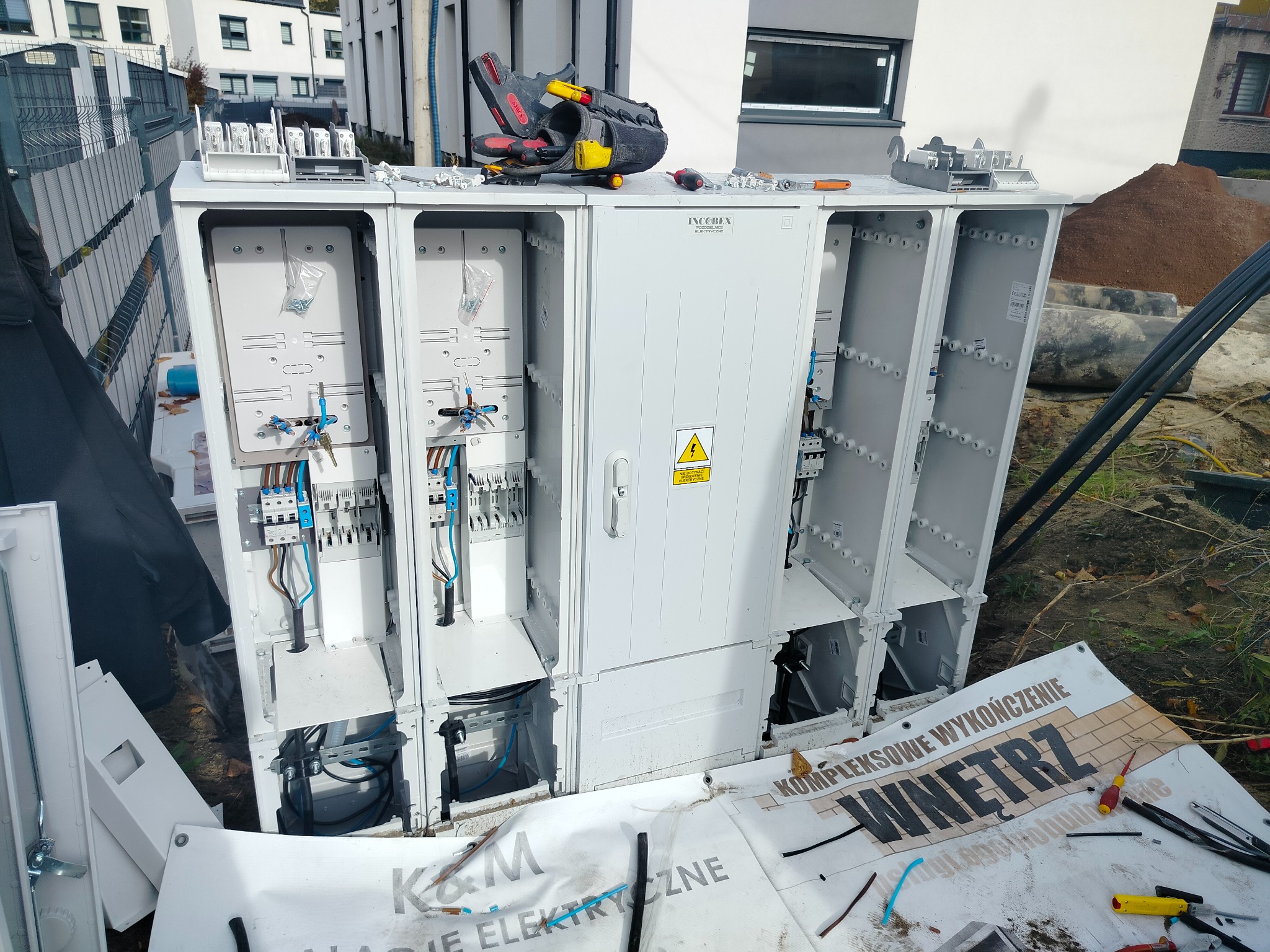 Otwarta, biała skrzynka elektryczna w trakcie montażu na zewnątrz budynku. Widoczne kable, narzędzia i elementy instalacji. Na górze torba narzędziowa.