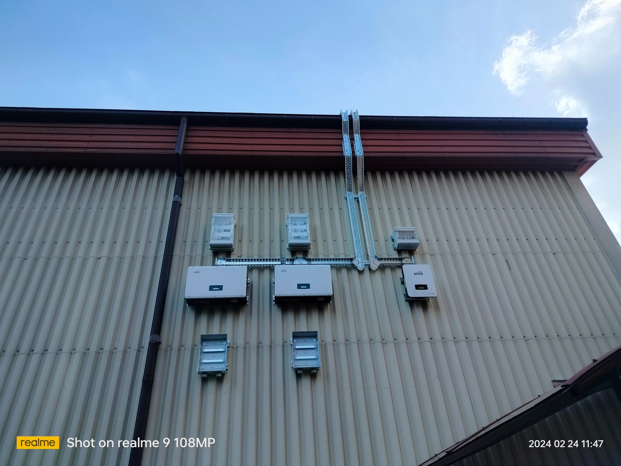 Elewacja budynku z zamontowanymi elementami instalacji fotowoltaicznej: falowniki i puszki przyłączeniowe, połączone metalowymi korytkami kablowymi.