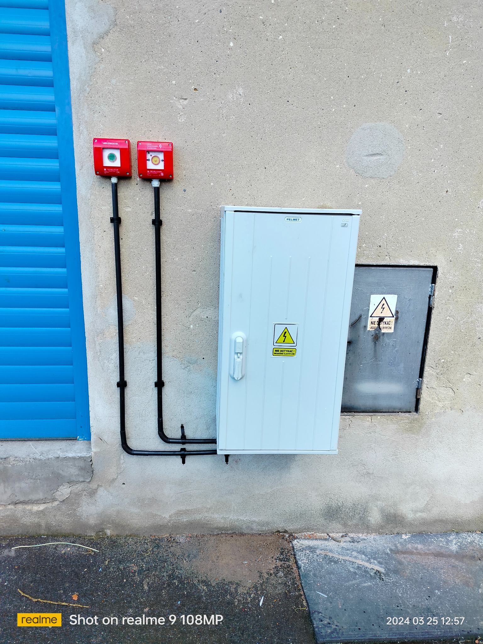 Zewnętrzna instalacja elektryczna: biała skrzynka PELMET z ostrzeżeniem, czerwone lampy sygnalizacyjne i niebieskie rolety na tle szarej ściany budynku.