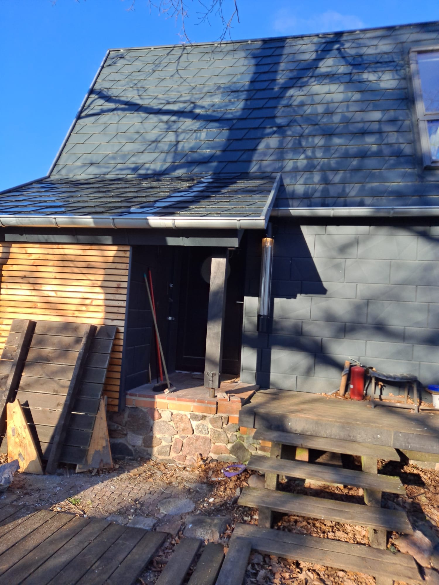Dom z dachem pokrytym szarą dachówką karpiówką, widoczne orynnowanie, fragment ściany wykończony drewnem i kamieniem, przed wejściem drewniane schody i narzędzia.