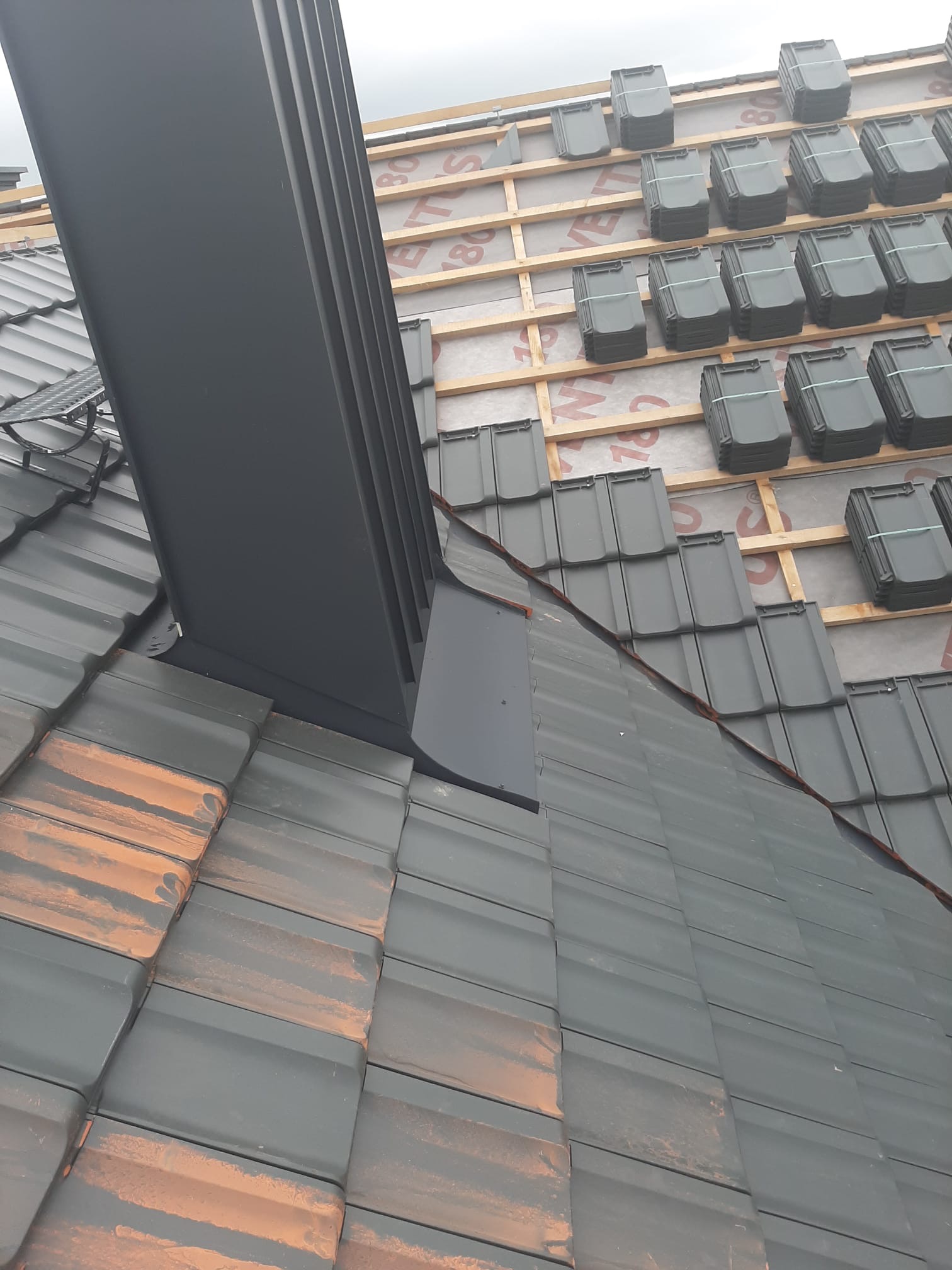 Montaż komina systemowego na dachu z dachówki ceramicznej, widoczne pakiety dachówek przygotowane do ułożenia na łatach.