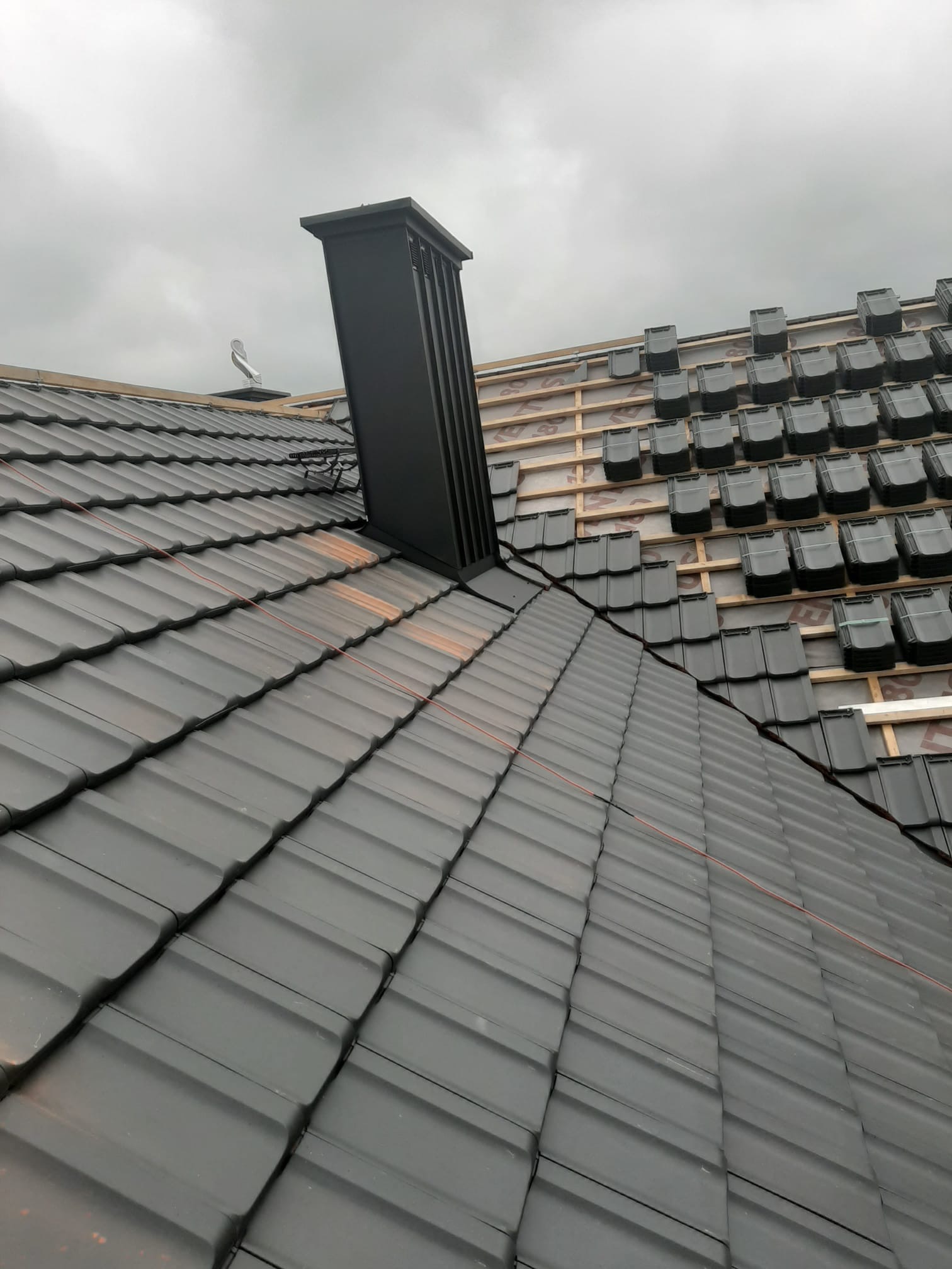 Częściowo pokryty dach z czarną dachówką ceramiczną, widoczny komin i fragment więźby dachowej z membraną, w tle pochmurne niebo.