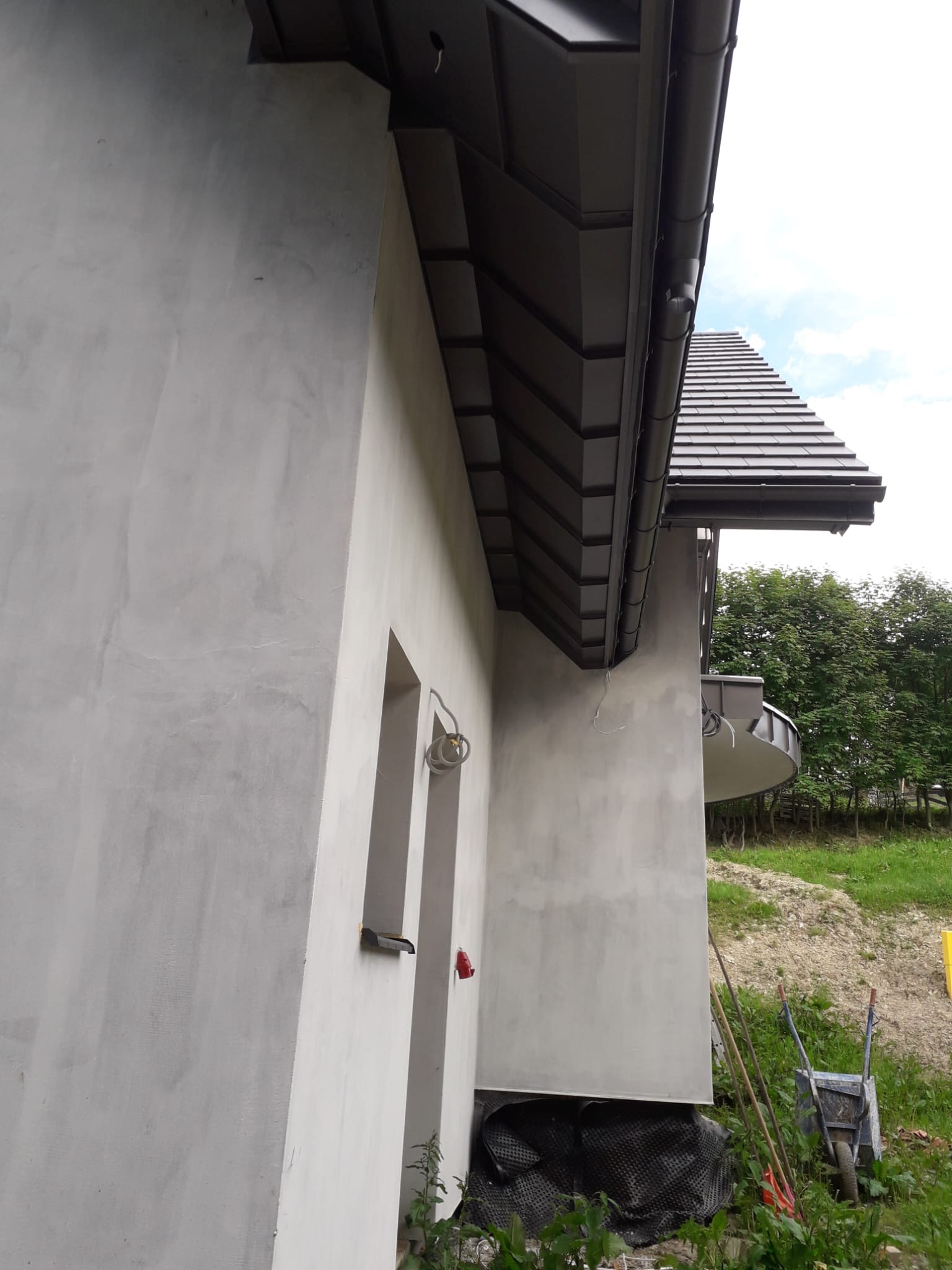 Szary budynek z ciemnoszarą dachówką i rynnami, widok z dołu, z widocznymi elementami wykończeniowymi i otoczeniem w postaci zieleni i wózka budowlanego.