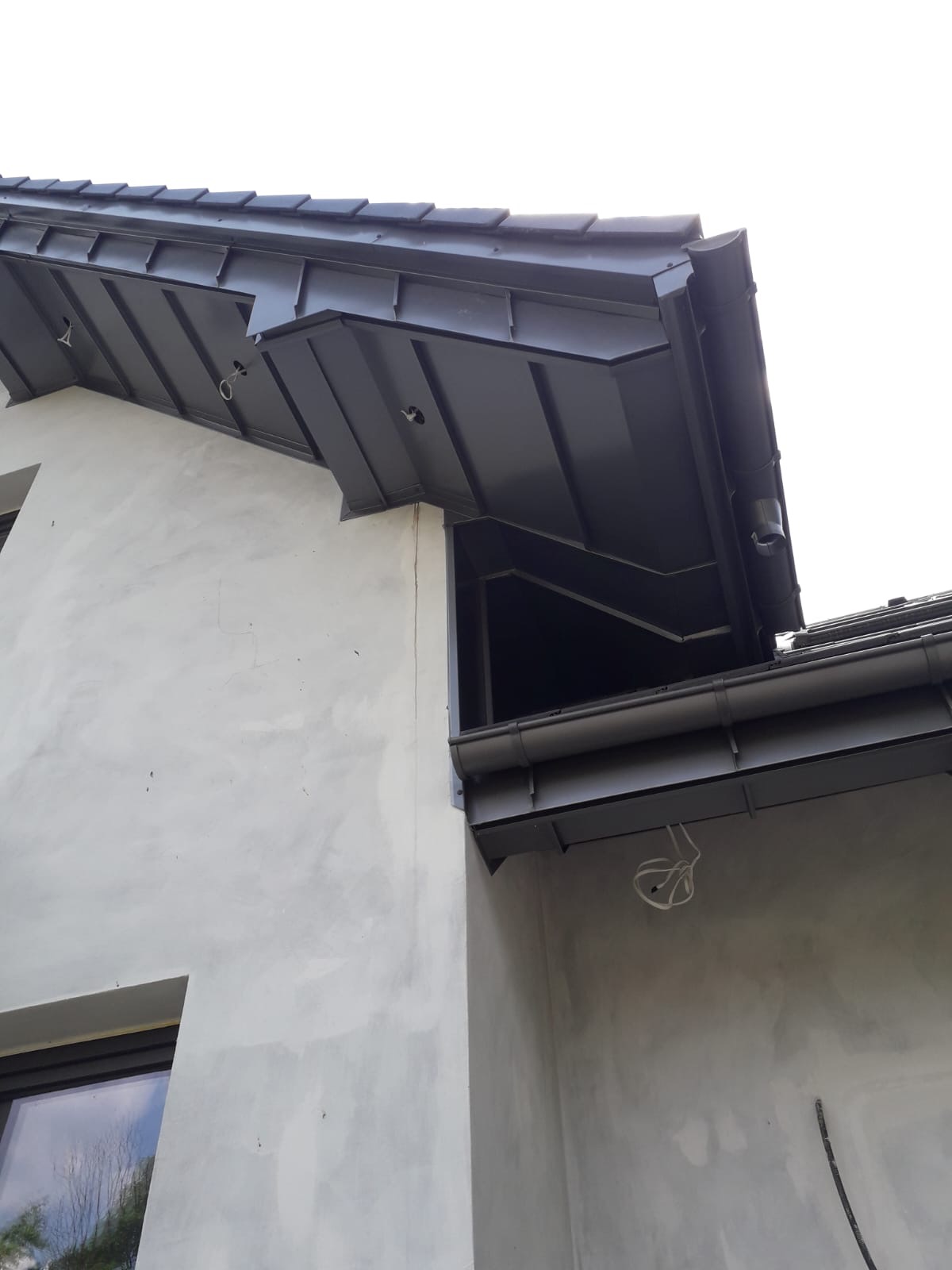 Szary metalowy dach z rynnami na budynku w trakcie wykończenia, widoczne wystające kable i surowa elewacja.