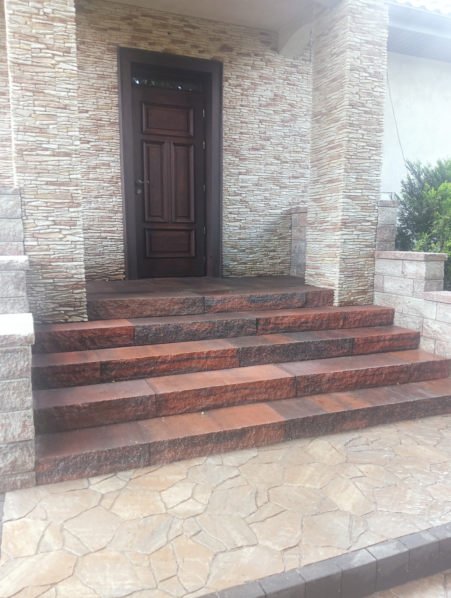 Wejście do domu z kamiennymi schodami w odcieniach brązu i beżu, elewacja budynku wykończona kamieniem imitującym cegłę, podłoże wyłożone nieregularnymi płytami.