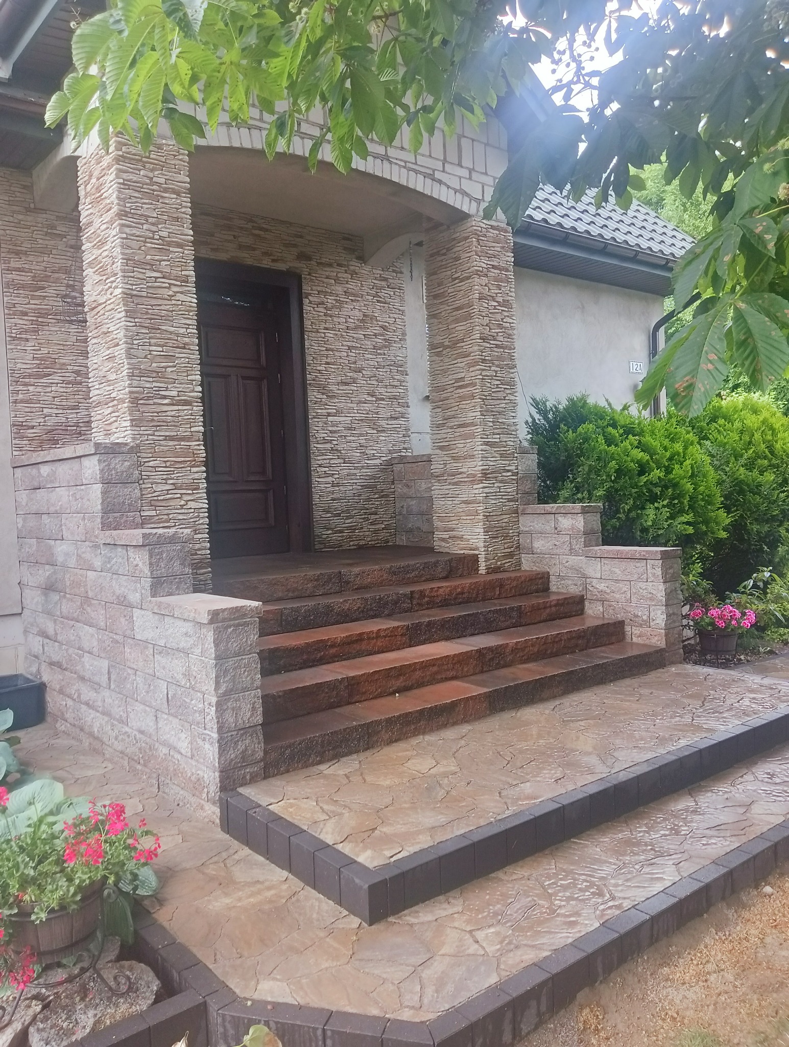 Wejście do domu z kamiennymi schodami i wybrukowanym tarasem, otoczone zielenią i kwiatami w donicach.