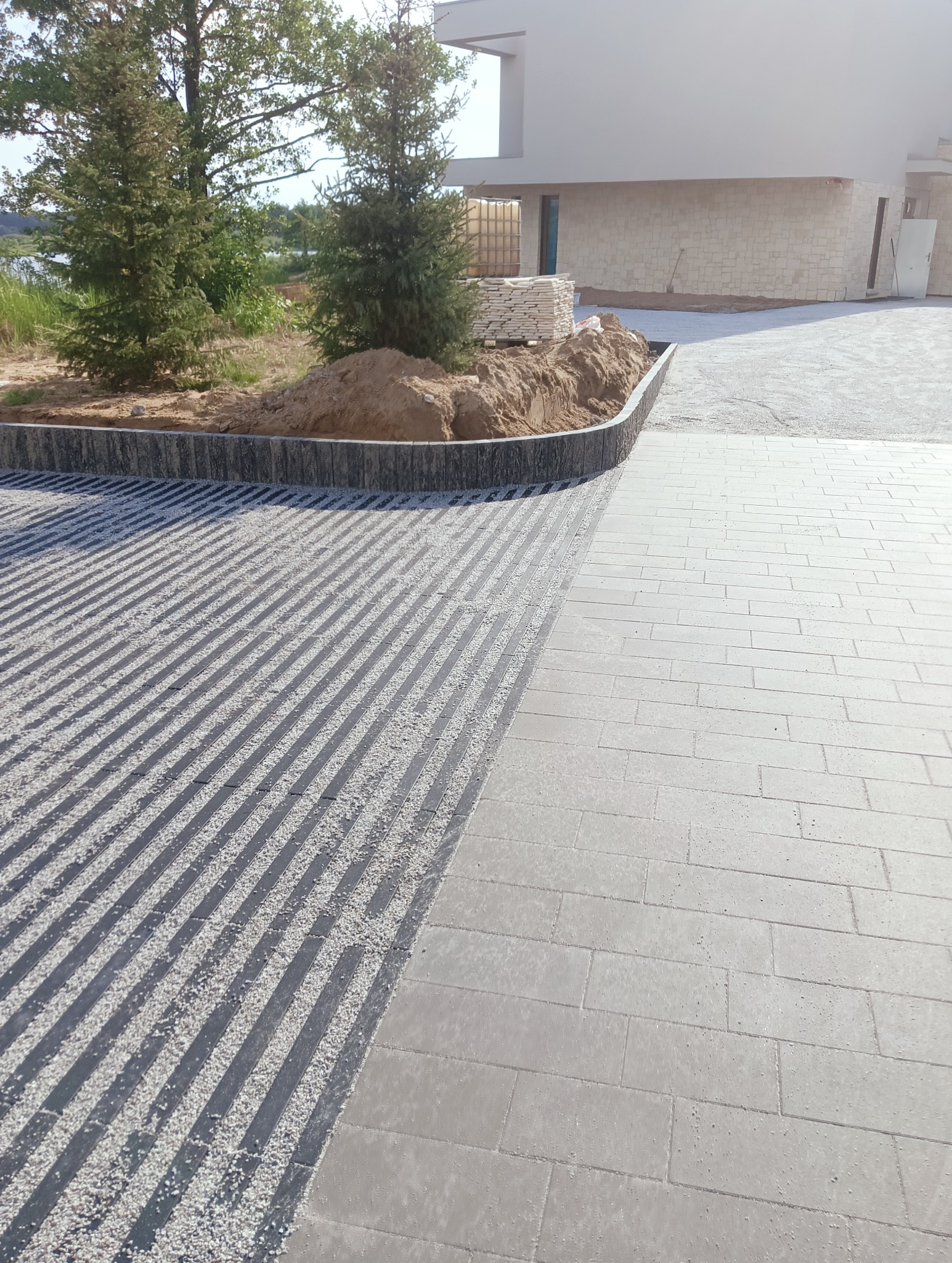 Nowoczesny podjazd z jasnoszarej kostki brukowej, część powierzchni z czarnymi pasami wypełnionymi drobnym białym grysem, obrzeże z ciemnych, pionowo ułożonych elementów, w tle trawnik, drzewa...