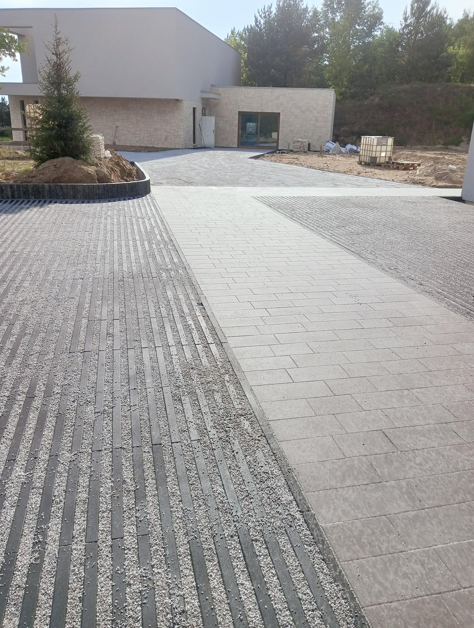 Ułożona nawierzchnia z dwóch rodzajów kostki: prostokątnej szarej kostki oraz kostki w formie podłużnych pasów wypełnionych drobnym kamieniem, na tle nowoczesnego domu.