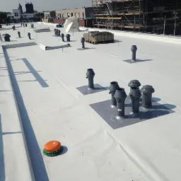 Biała, płaska powierzchnia dachu z widocznymi elementami wentylacyjnymi i zabezpieczeniami, w tle budynki i rusztowania.