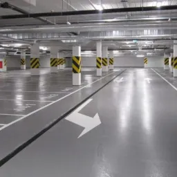 Gładka, szara posadzka przemysłowa z białymi oznaczeniami miejsc parkingowych i kierunkowymi strzałkami w hali garażowej, kolumny w pasy ostrzegawcze.