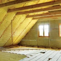 Poddasze w trakcie ocieplania wełną mineralną, widoczne drewniane krokwie, częściowo ułożona podłoga z desek i dwa okna, jedno otwarte, kabel zasilający na podłodze.