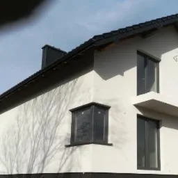 Nowoczesny dom jednorodzinny z białą elewacją, czarnym dachem i oknami w grafitowych ramach; widoczne ślady cieni drzew na ścianie.