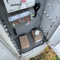 Otwarta szafka elektryczna z licznikiem MK-9z GSM/GPRS, modułem z bezpiecznikami, dwoma kartonowymi pudełkami z opisami, anteną i okablowaniem. Widoczne plomby i naklejki z numerami seryjnymi.