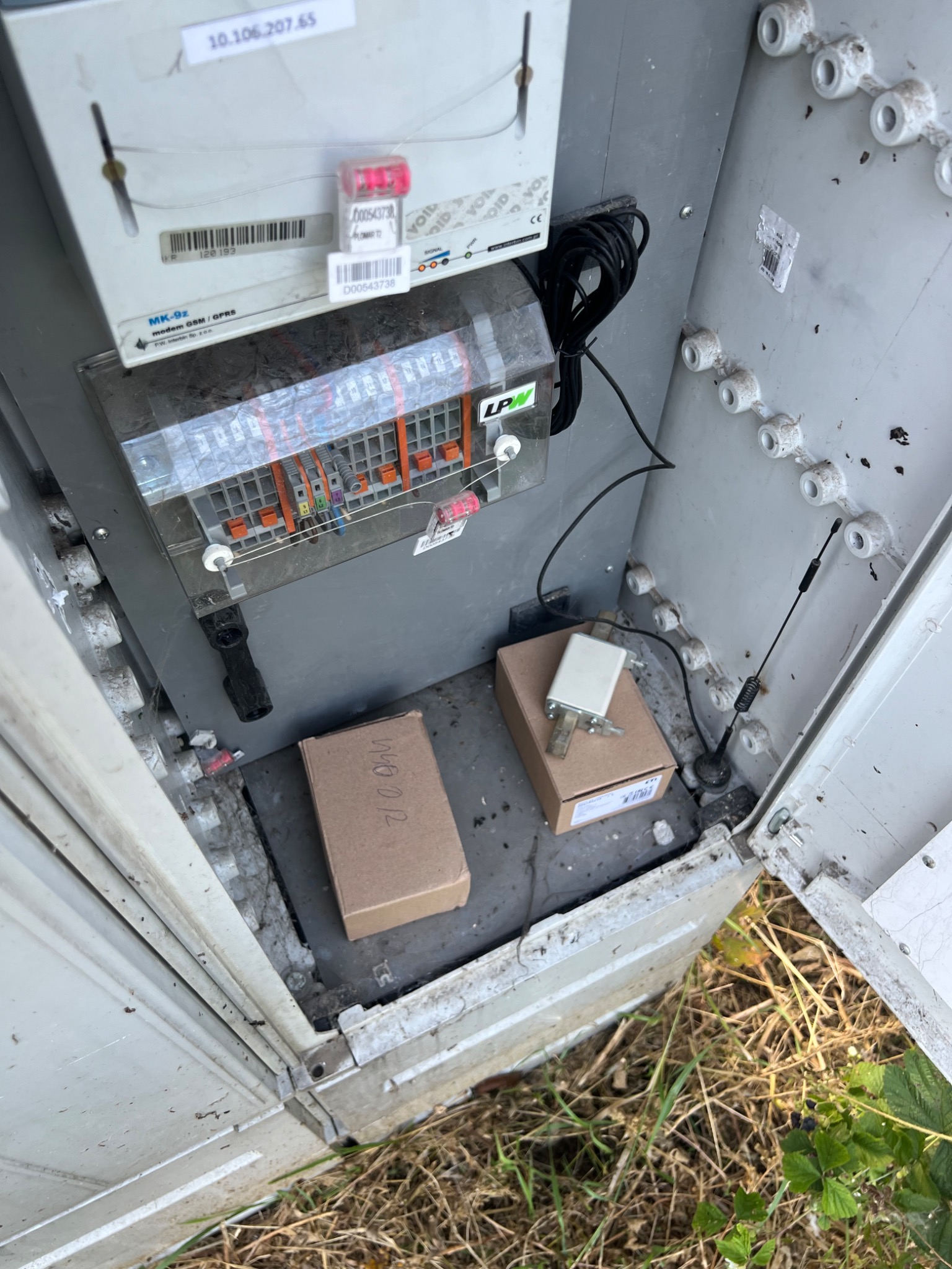 Otwarta szafka elektryczna z licznikiem MK-9z GSM/GPRS, modułem z bezpiecznikami, dwoma kartonowymi pudełkami z opisami, anteną i okablowaniem. Widoczne plomby i naklejki z numerami seryjnymi.