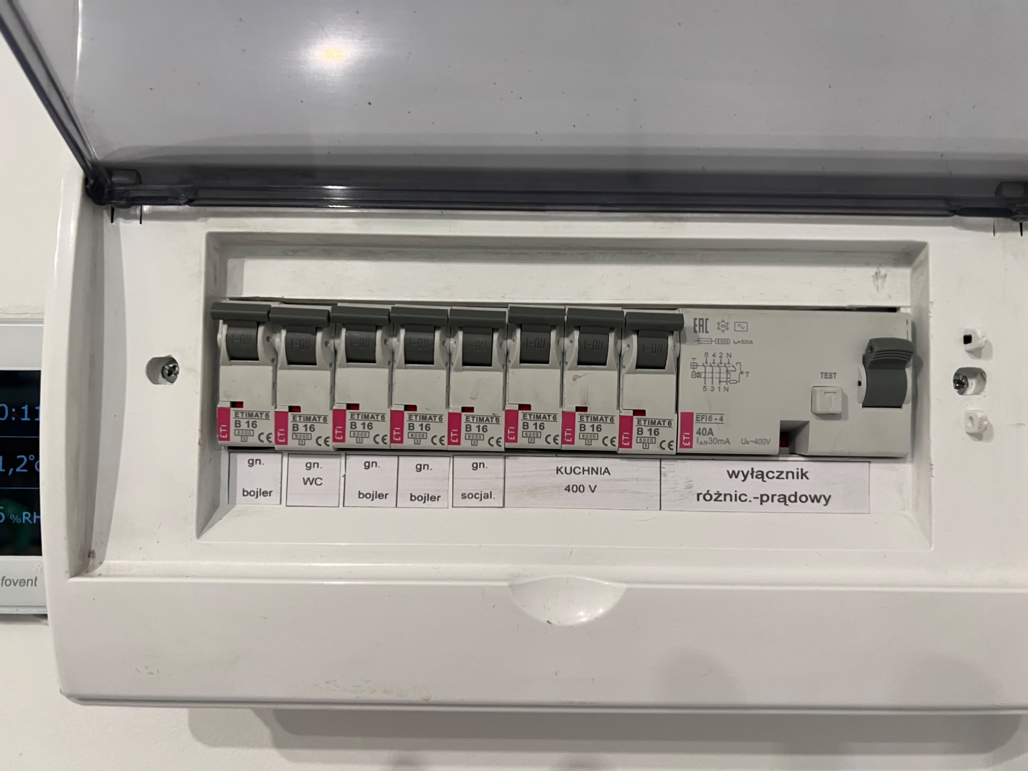 Otwarta skrzynka rozdzielcza z bezpiecznikami ETIMAT B16 opisana etykietami: bojler, WC, kuchnia 400V, wyłącznik różnicowoprądowy. Widoczny także wyłącznik główny oraz parametry techniczne.