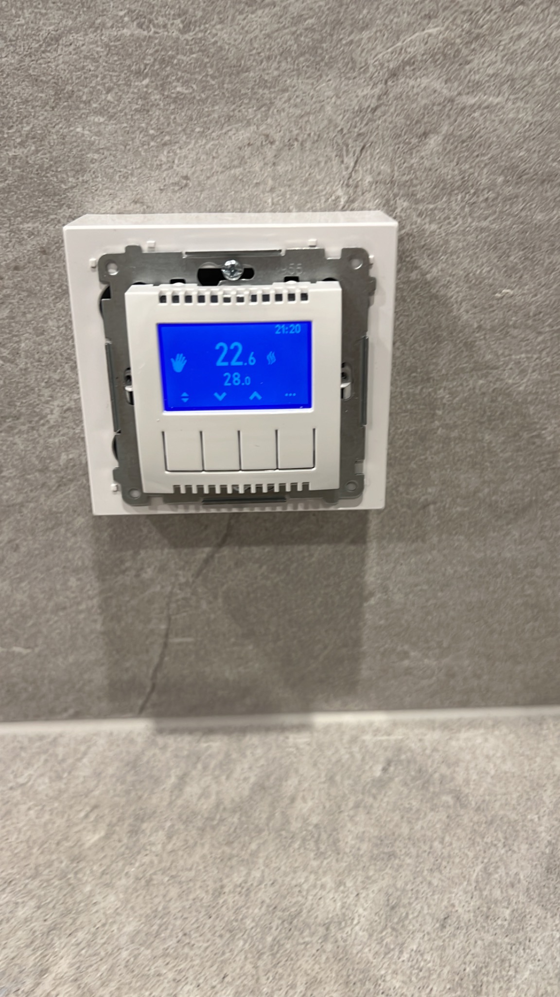 Biały panel sterowania ogrzewaniem podłogowym z niebieskim wyświetlaczem LCD, zamontowany na szarej ścianie z efektem betonu.