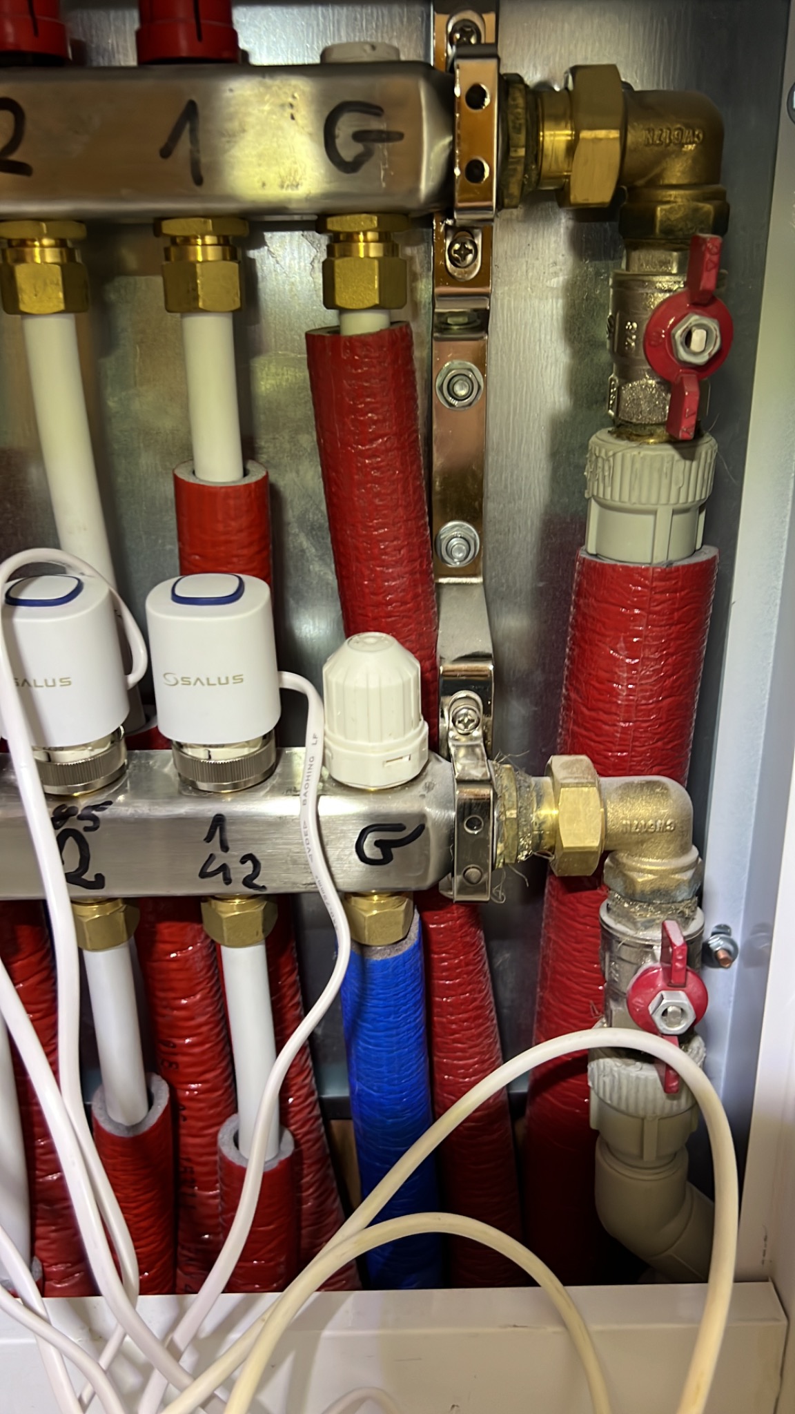 Rozdzielacz ogrzewania podłogowego z termostatami Salus, rury w izolacji czerwonej i niebieskiej, widoczne zawory i przewody.