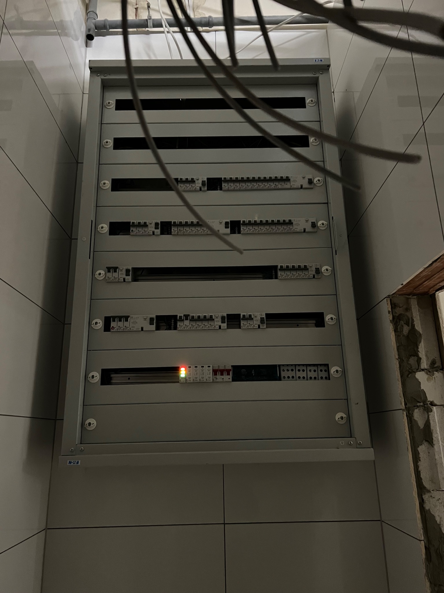 Szara skrzynka elektryczna z widocznymi bezpiecznikami i przewodami na tle białych płytek i surowego muru